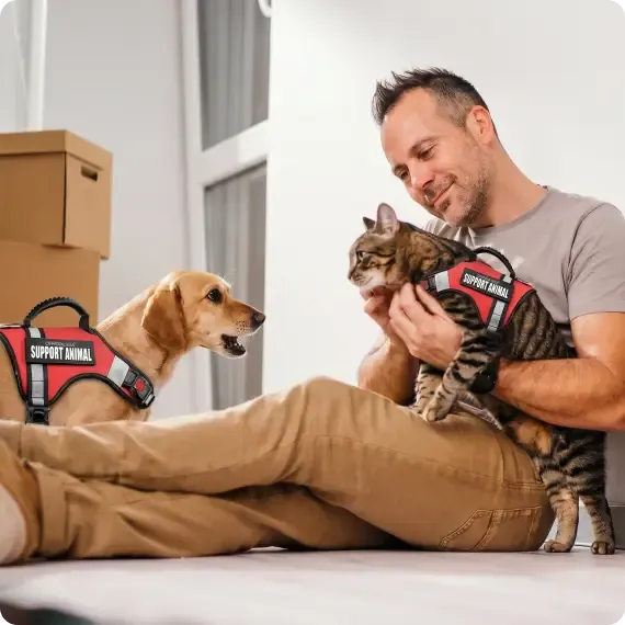 A man seated on the floor with his support dog and support cat, both wearing red Support Animal vests
